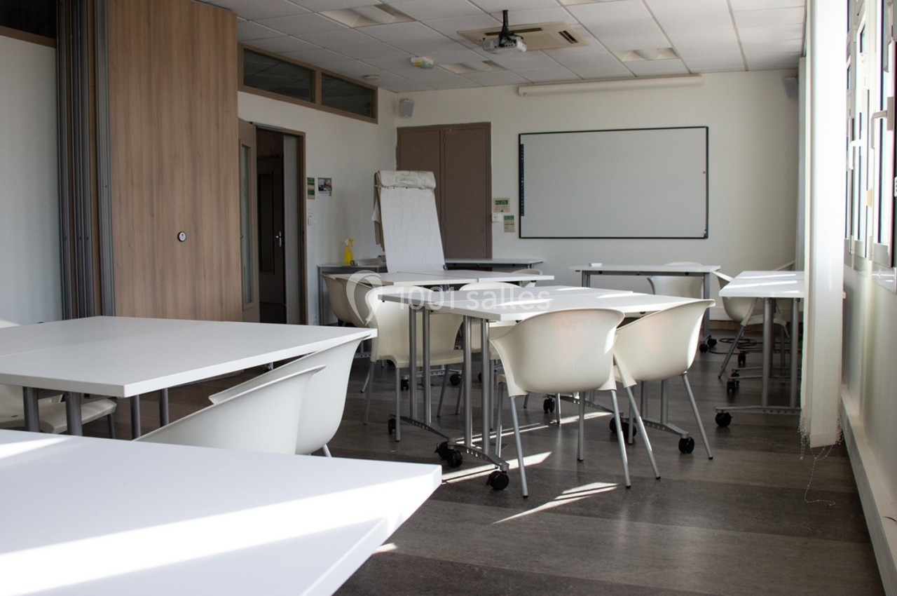 Salle de classe lumineuse avec des tables blanches, des chaises modernes et un tableau blanc au mur.