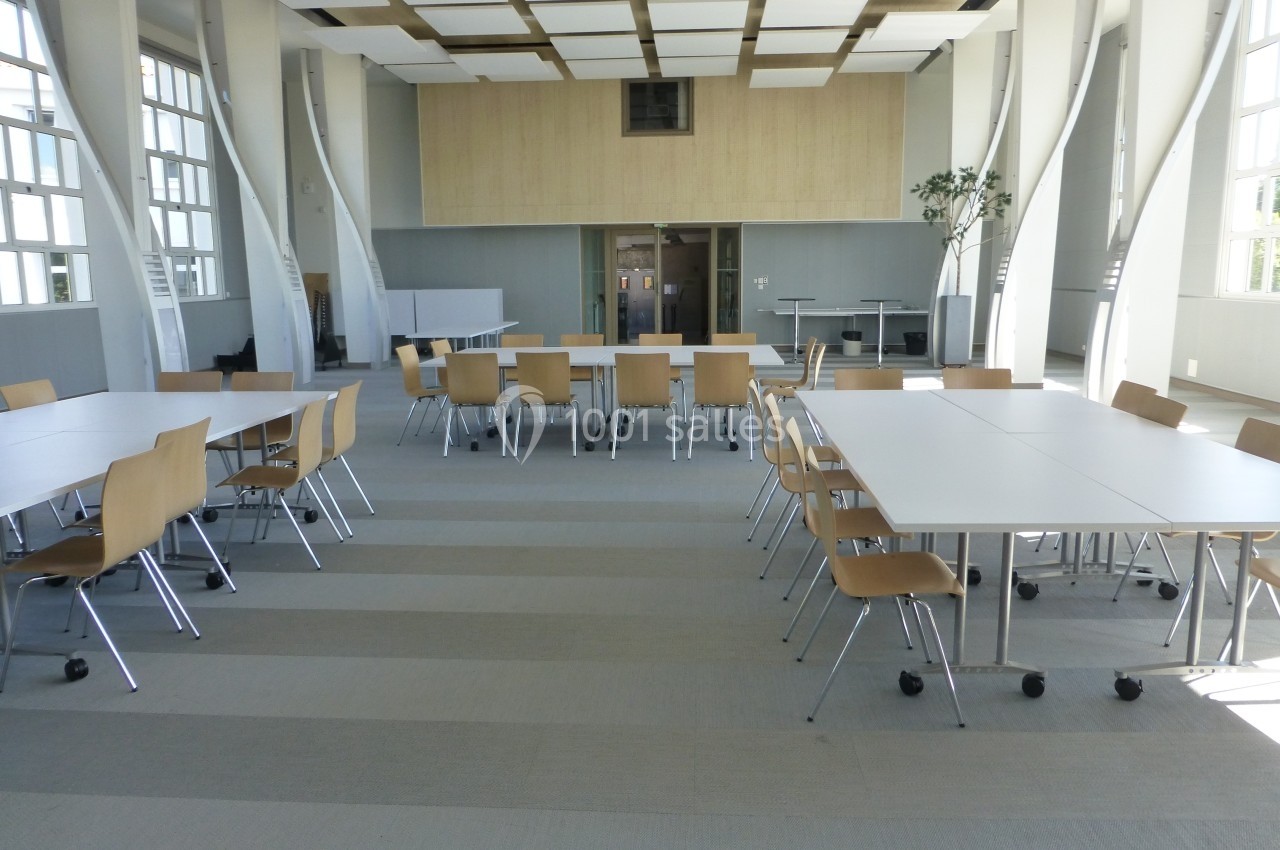 Salle lumineuse avec de grandes fenêtres, tables blanches et chaises en bois disposées pour une réunion ou un travail…