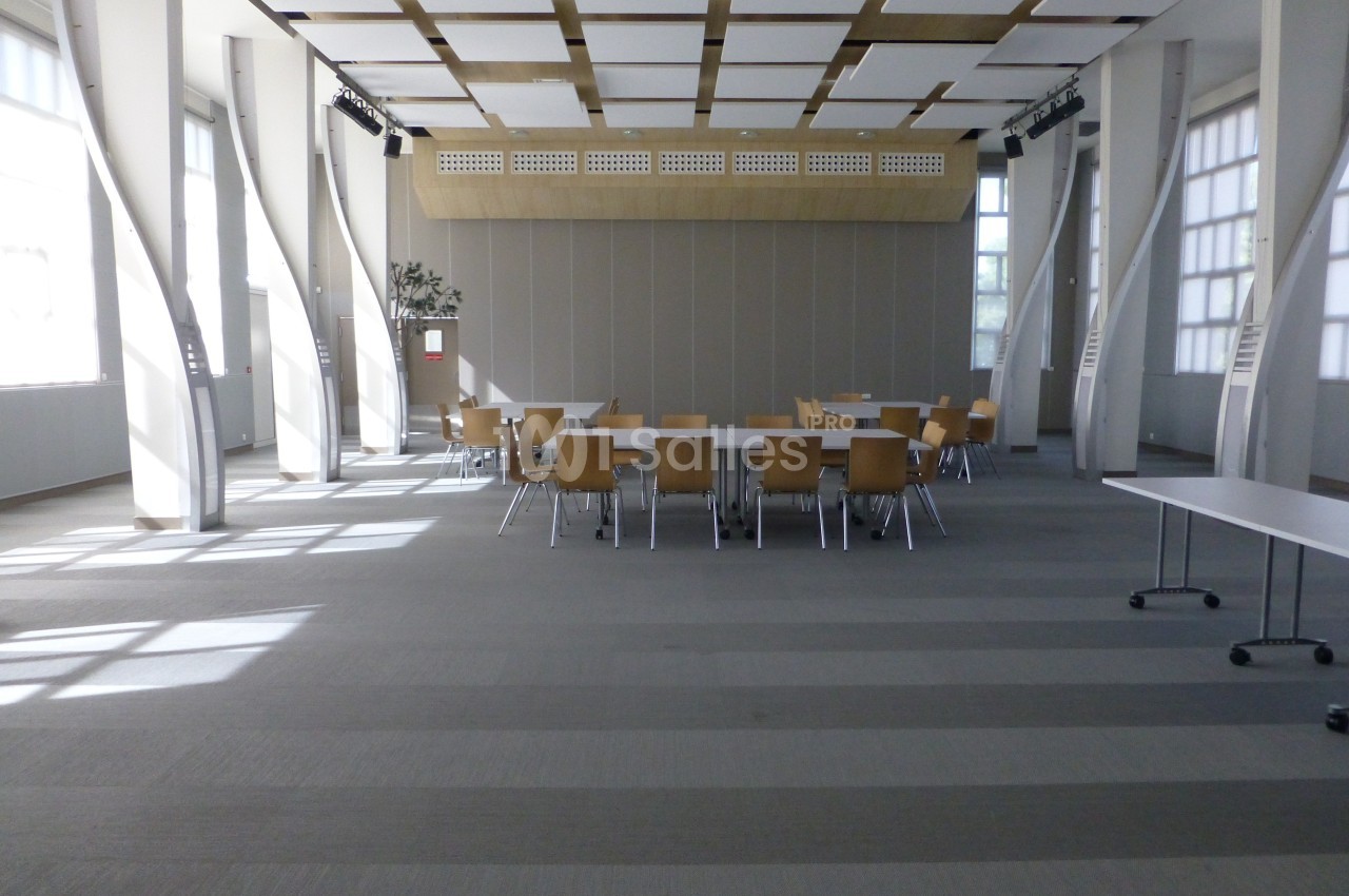 Salle de réunion lumineuse avec des tables et des chaises en bois disposées au centre, entourées de grandes fenêtres.