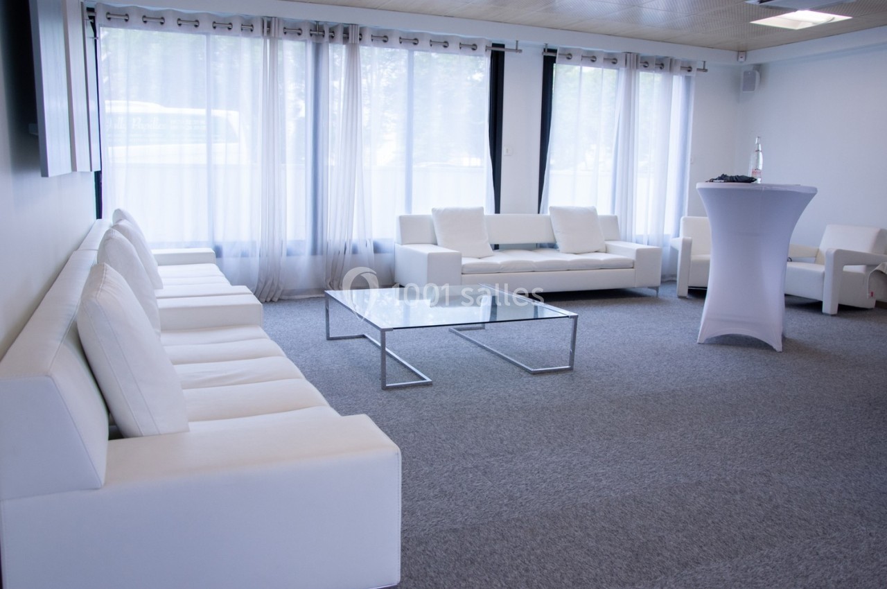 Salle lumineuse avec canapés blancs, table basse en verre et grandes fenêtres avec rideaux translucides.