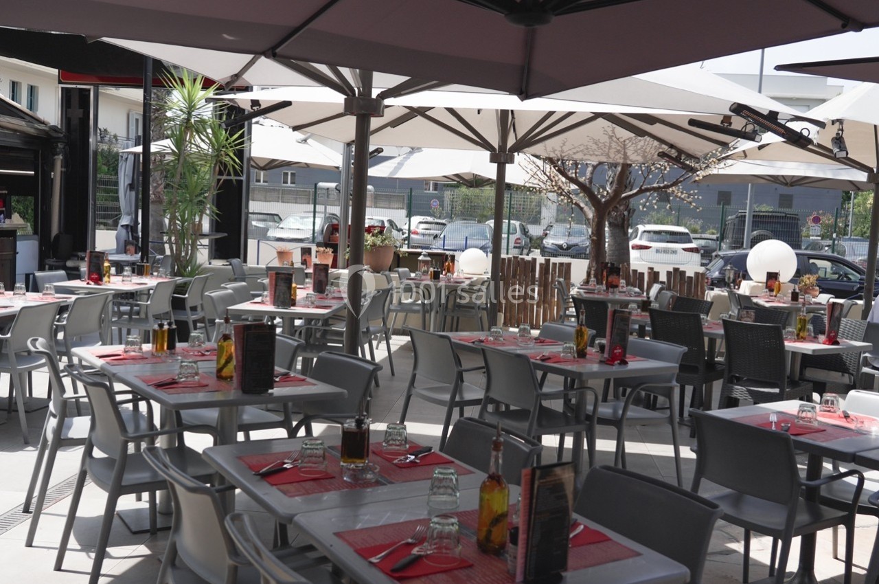Location salle Villeneuve-Loubet (Alpes-Maritimes) - Pizzatti Villeneuve Loubet #6 Terrasse de restaurant avec tables dressées, chaises grises, parasols ouverts et vue sur un parking en arrière-plan.