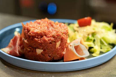 Assiette contenant un tartare de viande, des tranches de jambon cru et une salade verte avec une tomate cerise.