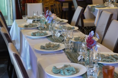Table dressée avec nappes blanches, serviettes bleues, verres, couverts et décorations florales colorées.