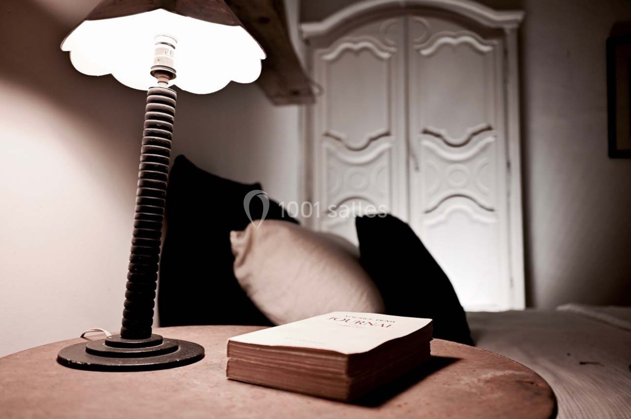 Lampe allumée sur une table de chevet en bois avec un livre, lit et armoire en arrière-plan.