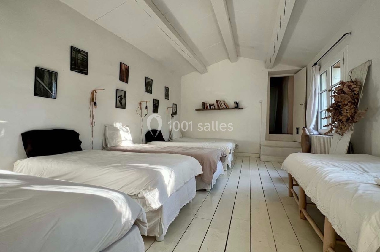 Quatre lits simples alignés dans une chambre lumineuse avec murs blancs, décor minimaliste et poutres apparentes.
