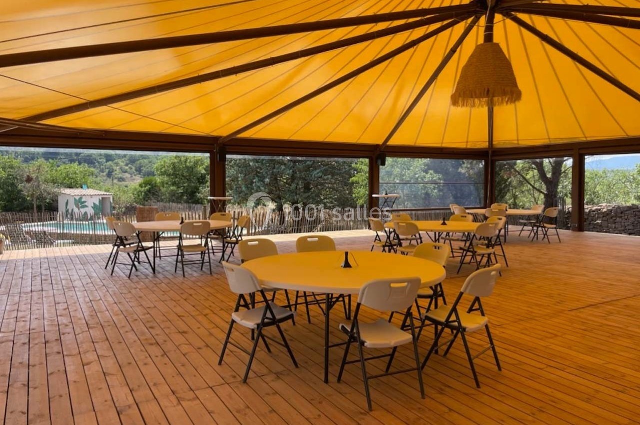 Espace couvert avec tables et chaises sur plancher en bois, entouré de nature et vue sur une piscine.
