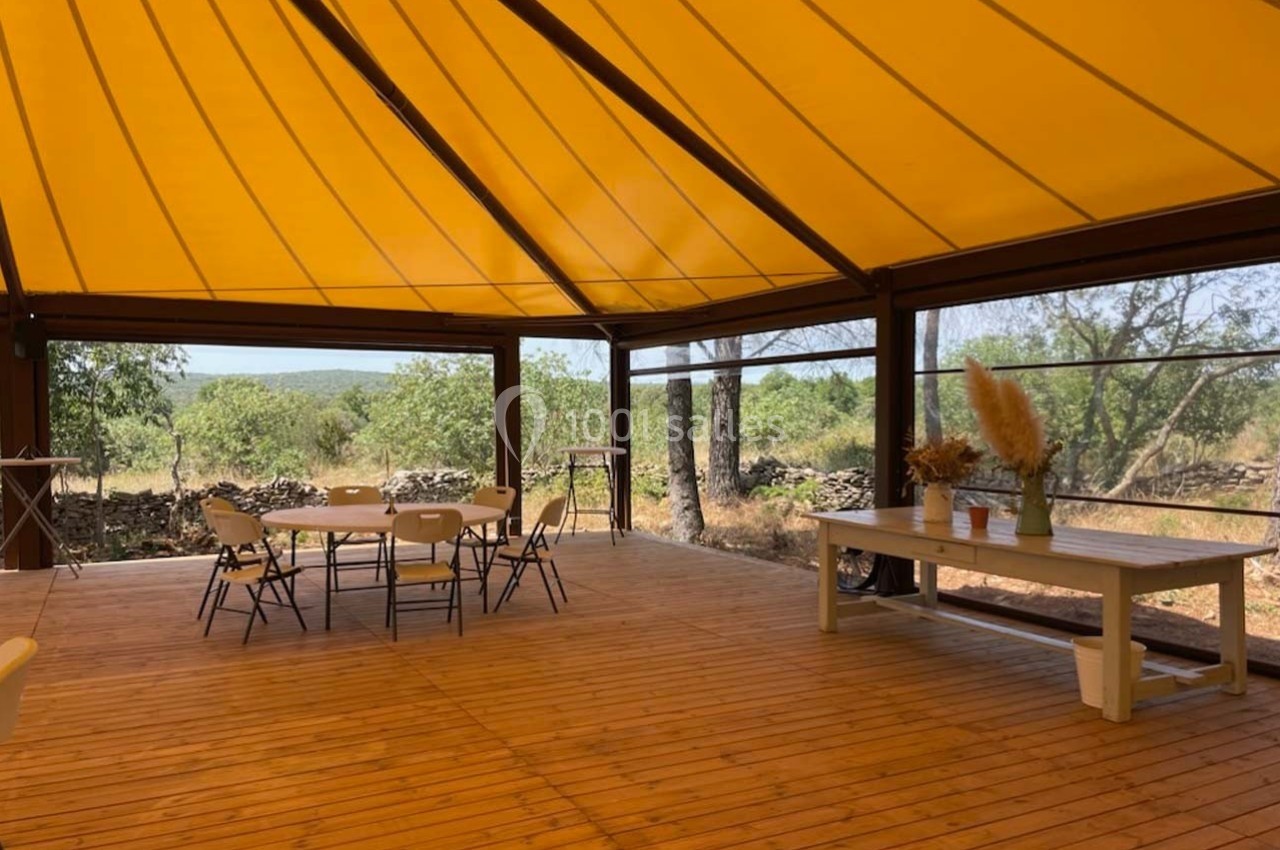 Terrasse en bois couverte d'une toile jaune, avec des tables et chaises, entourée de végétation.