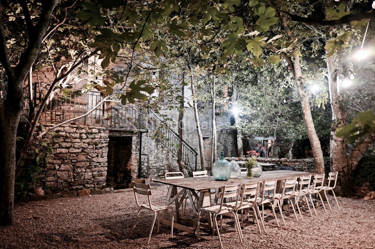 Grande table en bois entourée de chaises dans une cour extérieure éclairée, entourée de murs en pierre et d'arbres.