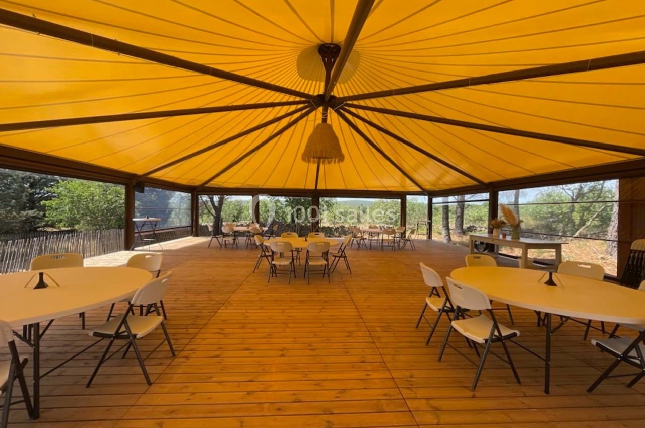 Salle extérieure couverte avec toit jaune, tables rondes et chaises pliantes sur un plancher en bois.