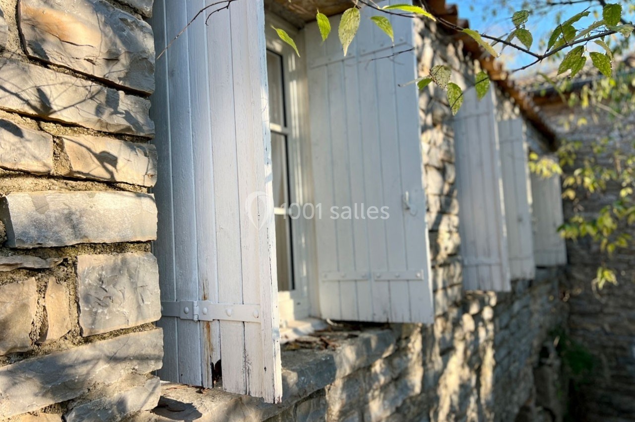 Façade en pierre avec des volets en bois blancs ouverts, éclairée par une lumière naturelle et entourée de végétation.