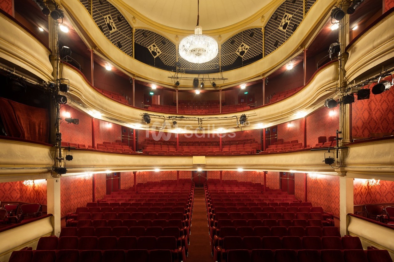 Salle de théâtre vide avec sièges rouges, balcons ornés et un grand lustre central suspendu au plafond.