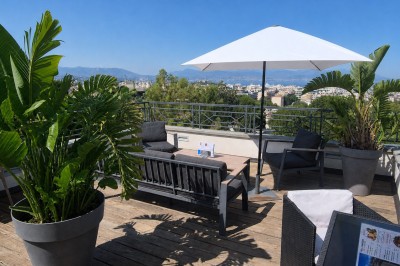 Terrasse ensoleillée avec mobilier de jardin, parasol blanc et plantes en pot, offrant une vue sur une ville et des collines.