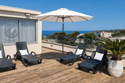 Terrasse ensoleillée avec mobilier de jardin, parasol blanc et plantes en pot, offrant une vue sur une ville et des collines.