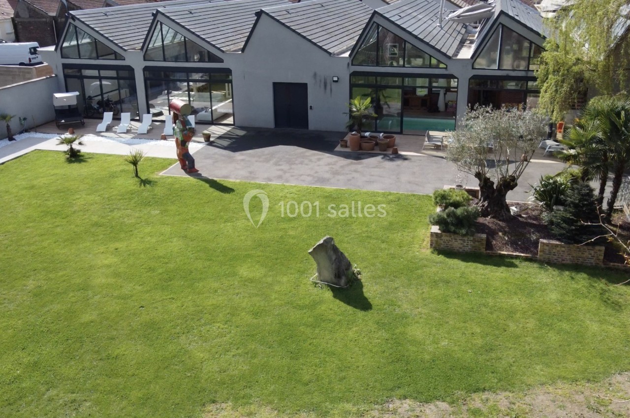 Vue d'un grand jardin avec pelouse, sculptures, olivier, et une maison moderne aux larges baies vitrées.