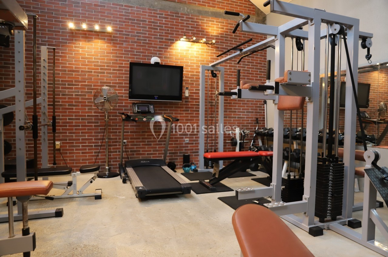 Salle de sport équipée avec tapis de course, machines de musculation et mur en briques apparentes.