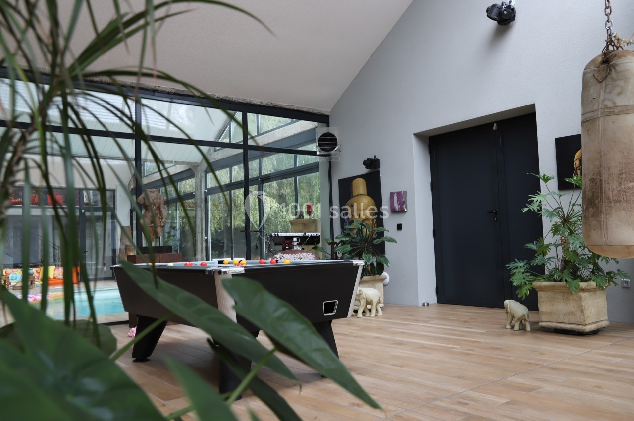 Salle lumineuse avec table de billard, plantes, sac de frappe et vue sur une piscine intérieure à travers des baies vitrées.