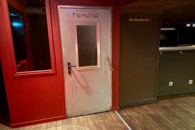 Bar en bois sombre avec comptoir équipé de crochets, étagères à verres et tableau noir affichant le menu.