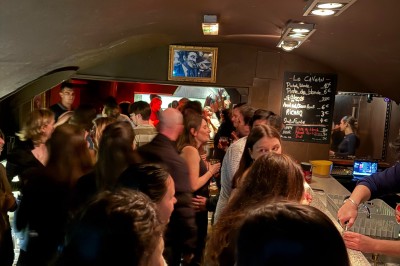 Bar en bois sombre avec comptoir équipé de crochets, étagères à verres et tableau noir affichant le menu.