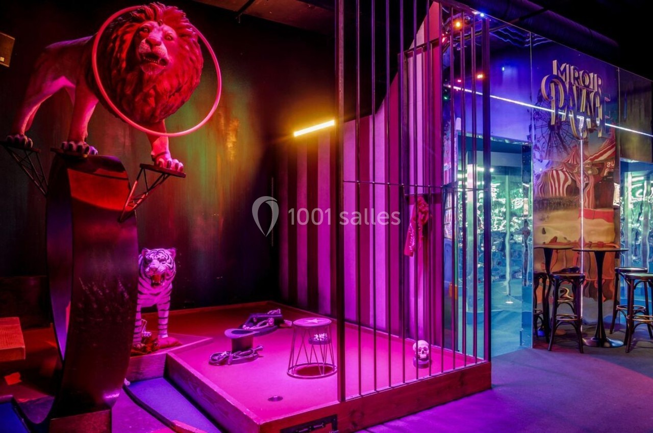 Décor de cirque avec lion sur un socle, tigre en cage, accessoires au sol et éclairage coloré.