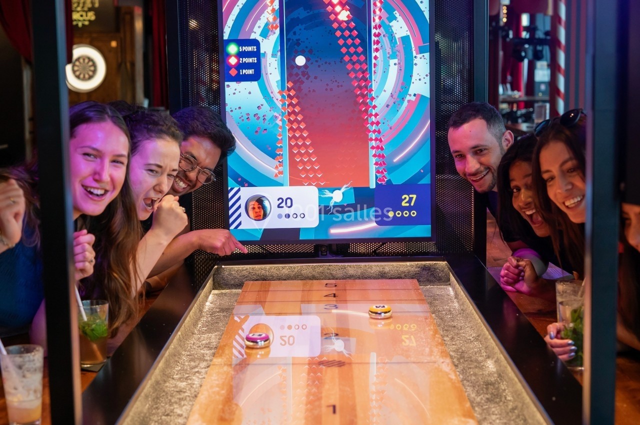 Des personnes jouent à un jeu interactif sur une table en bois avec un écran affichant les scores.