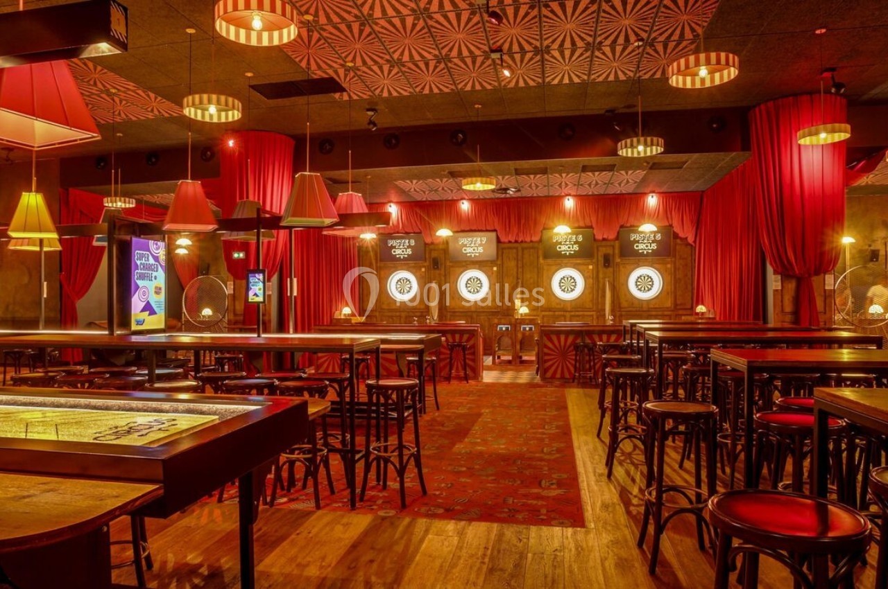 Salle de jeux avec cibles de fléchettes, tables hautes en bois, chaises et éclairage chaleureux sous des rideaux rouges.