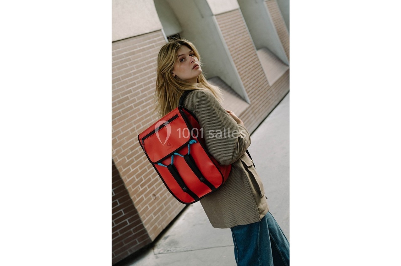 Femme portant un sac à dos rouge vif, marchant près d'un mur en briques dans un environnement urbain.