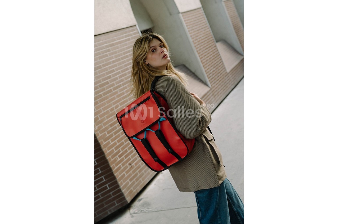 Femme portant un sac à dos rouge vif, marchant près d'un mur en briques dans un environnement urbain.
