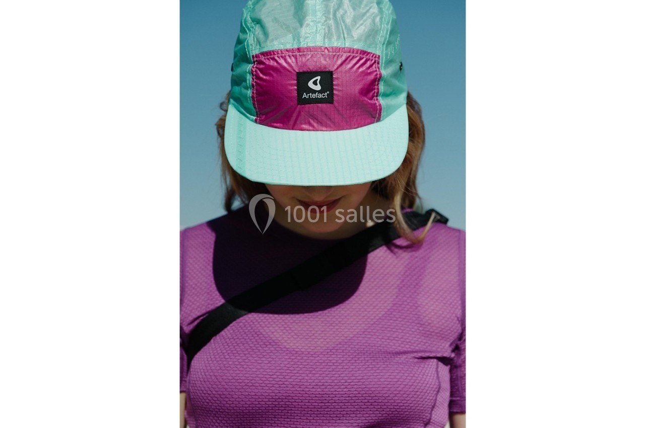 Femme portant une casquette bicolore turquoise et rose, avec un haut violet, visage partiellement caché.