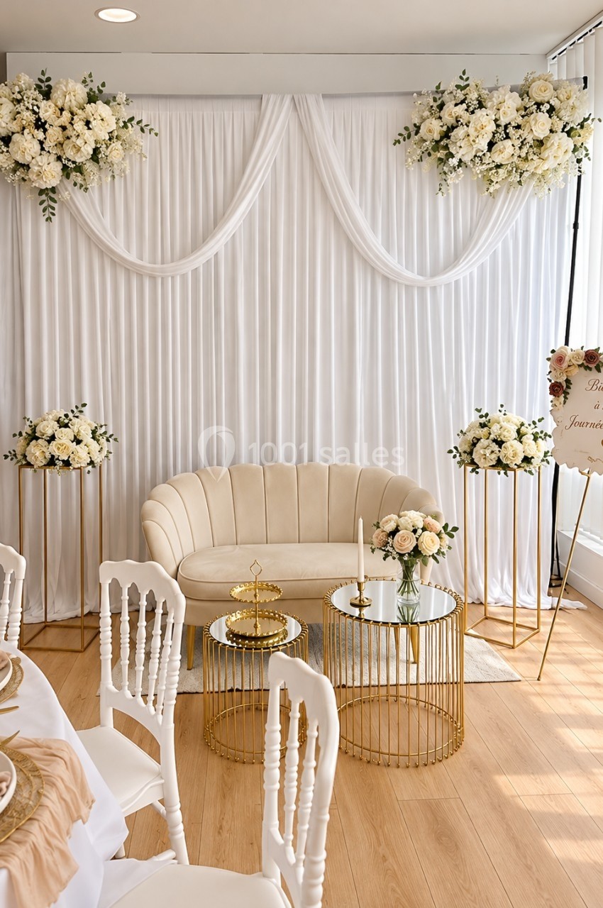 Décor élégant avec canapé beige, tables dorées, fleurs blanches et rideaux blancs dans une salle lumineuse.