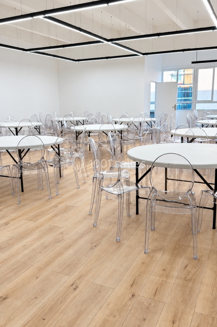 Salle lumineuse avec tables rondes blanches, chaises transparentes et sol en parquet clair.