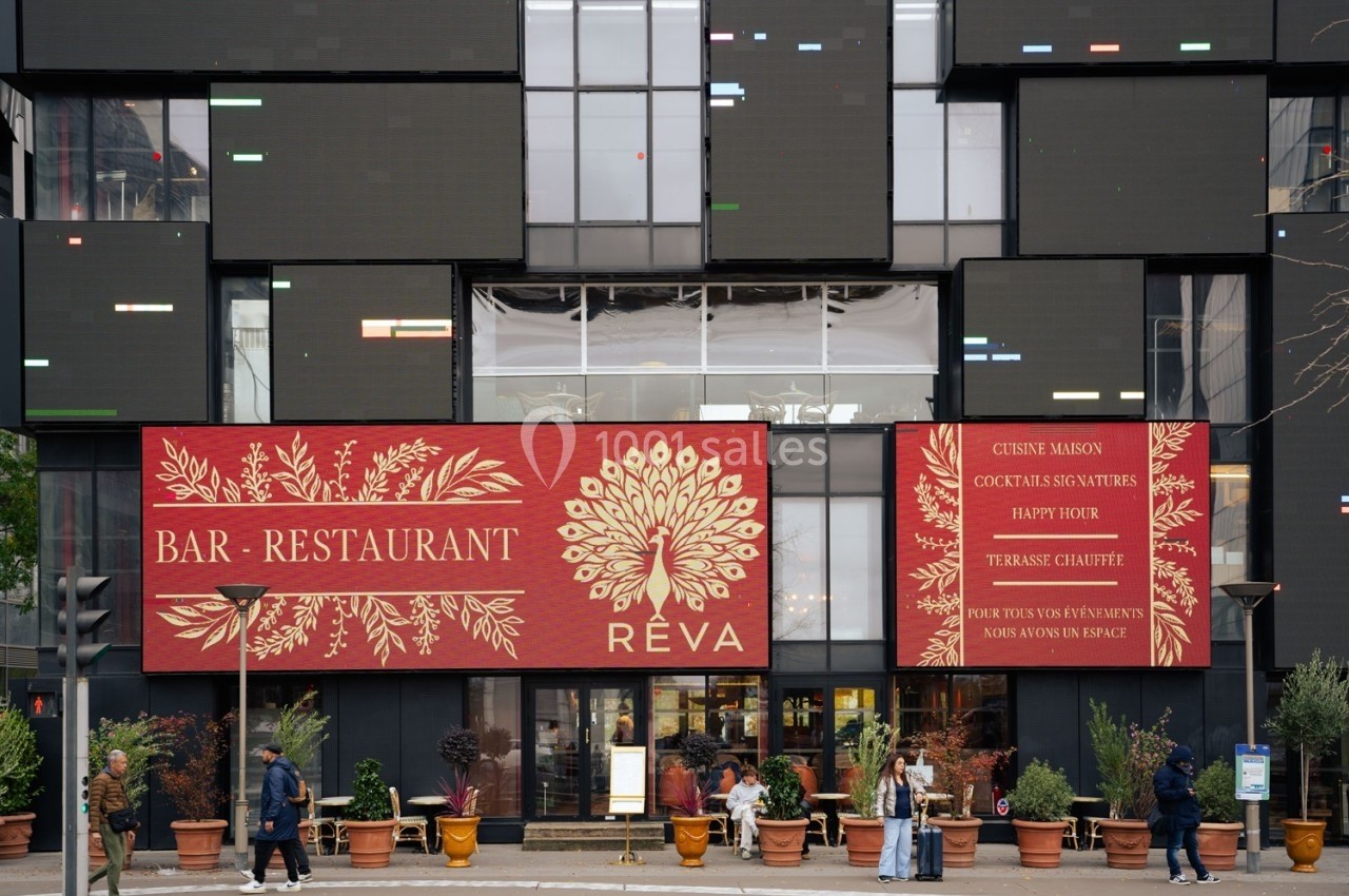 Façade d'un bar-restaurant avec terrasses, enseignes rouges et vitrines, entourée de plantes et de passants.