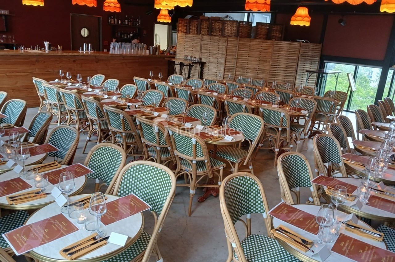 Salle de restaurant avec tables dressées, chaises en osier, nappes blanches et éclairage chaleureux par des suspensions…