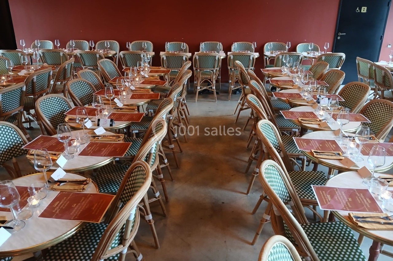 Salle de restaurant avec des tables dressées, nappes blanches, menus rouges et chaises en osier devant un mur rouge.