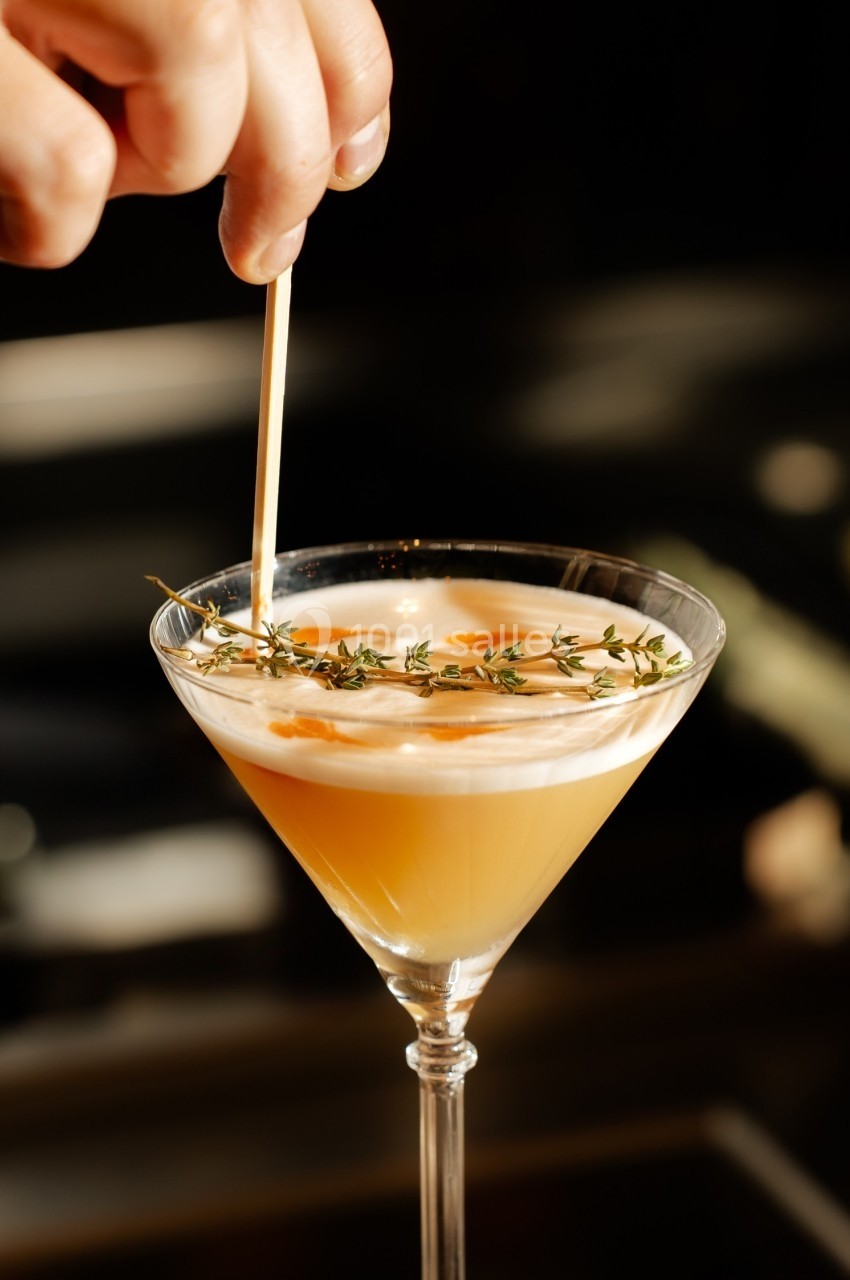 Un cocktail jaune dans un verre à martini décoré de thym et d'une mousse blanche, une main ajustant une garniture.