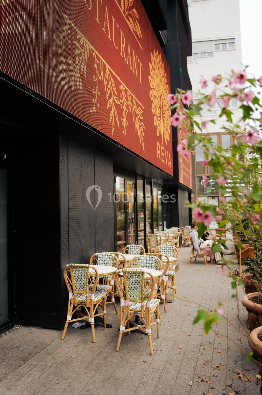 Terrasse d'un restaurant avec chaises en rotin, située sur un trottoir bordé de plantes fleuries.
