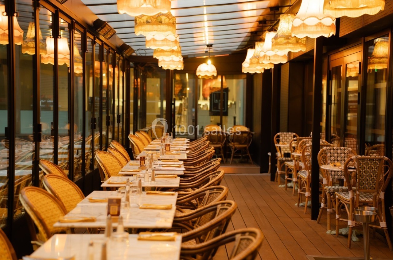 Terrasse couverte d'un restaurant avec tables dressées, chaises en rotin et éclairage chaleureux sous des lampes suspendues.