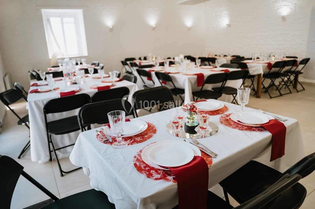 Salle décorée pour un repas, avec des tables dressées en blanc et rouge, chaises noires et lumière naturelle.