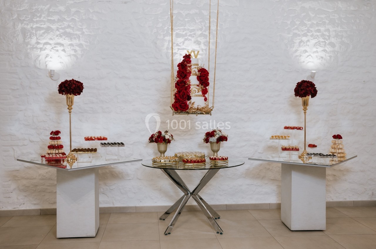 Table de desserts élégante avec gâteaux, macarons et fleurs rouges, disposée sur des tables en verre dans une salle blanche.