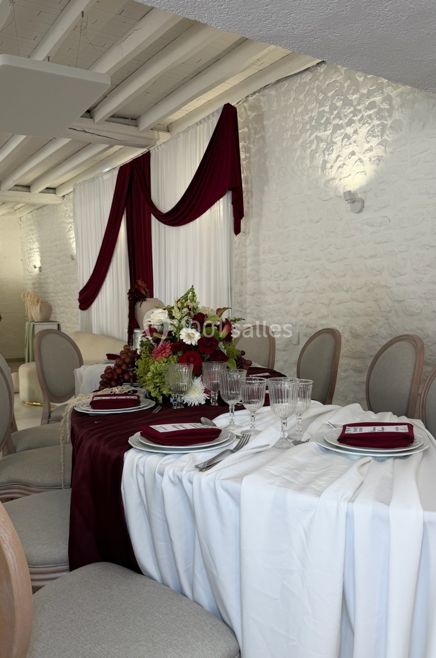 Table élégamment dressée avec nappes blanches et bordeaux, décorée de fleurs et entourée de chaises rembourrées.