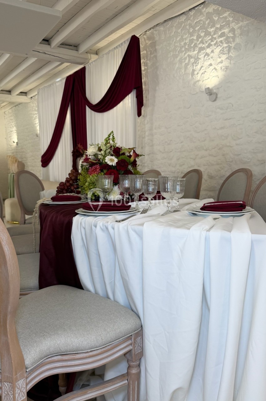 Table élégamment dressée avec nappes blanches, vaisselle raffinée et décoration florale rouge dans une salle lumineuse.