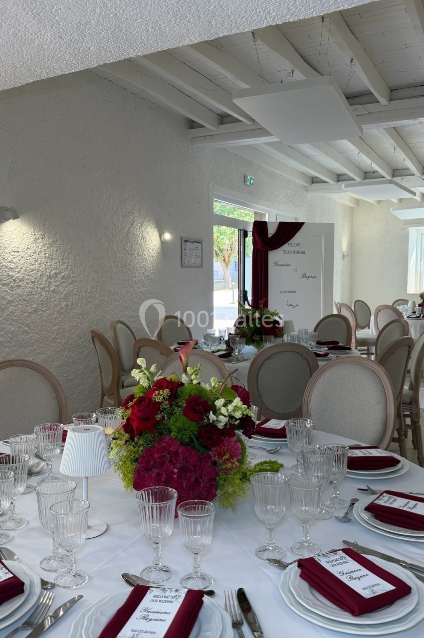 Salle de réception décorée avec des tables élégantes, nappes blanches, fleurs rouges et vertes, et serviettes bordeaux.