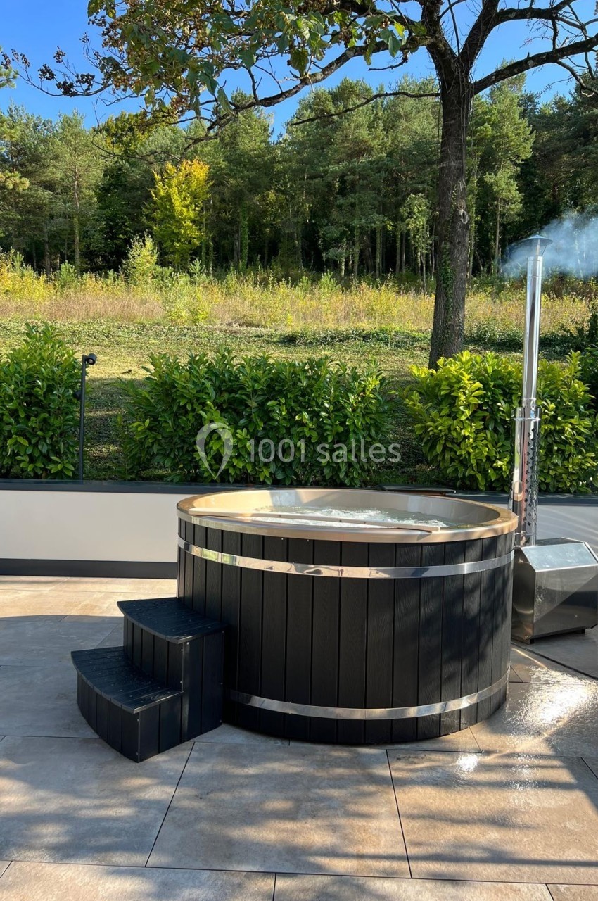 Bain nordique en bois sombre avec cheminée, installé sur une terrasse ensoleillée près d'une haie et d'une forêt.
