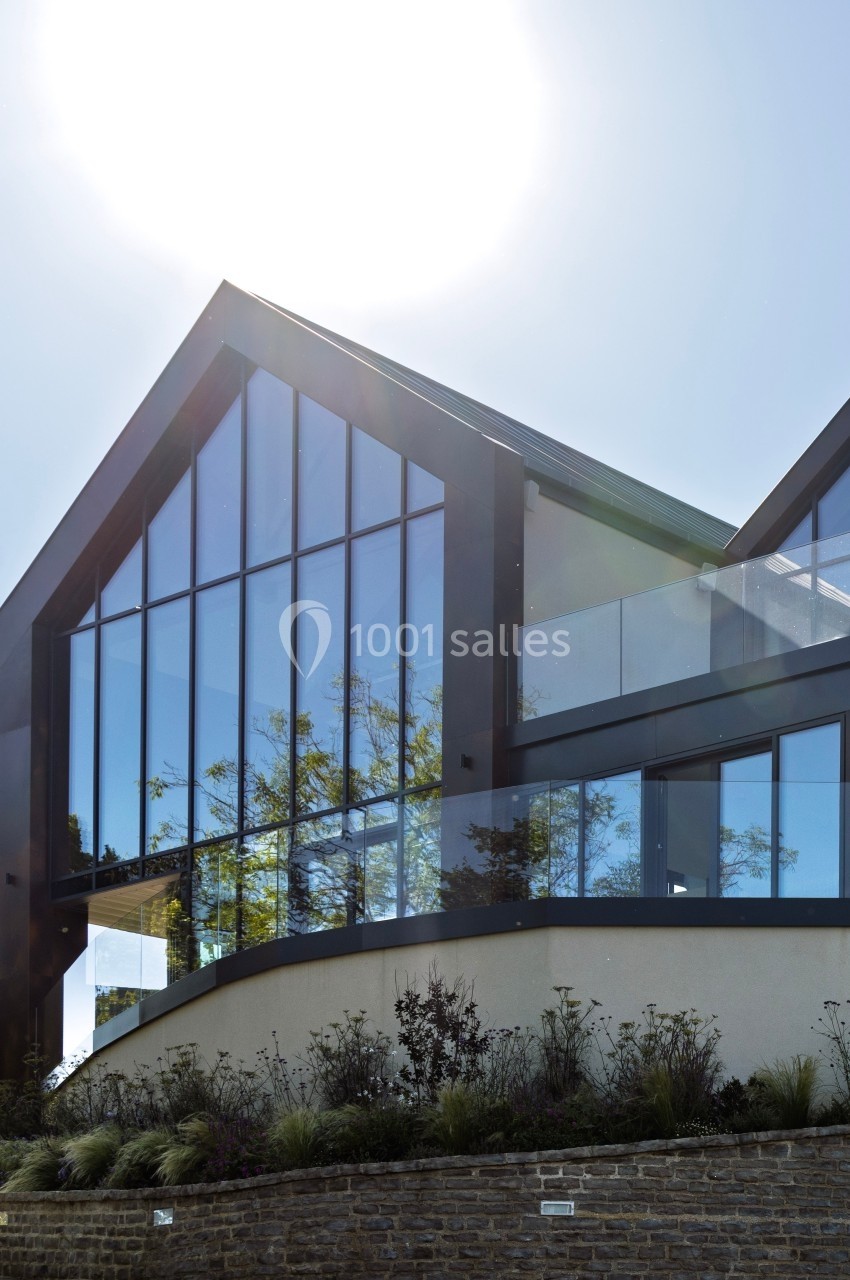 Façade moderne d'une maison avec de grandes baies vitrées reflétant le ciel, entourée de végétation et d'un mur en pierre.
