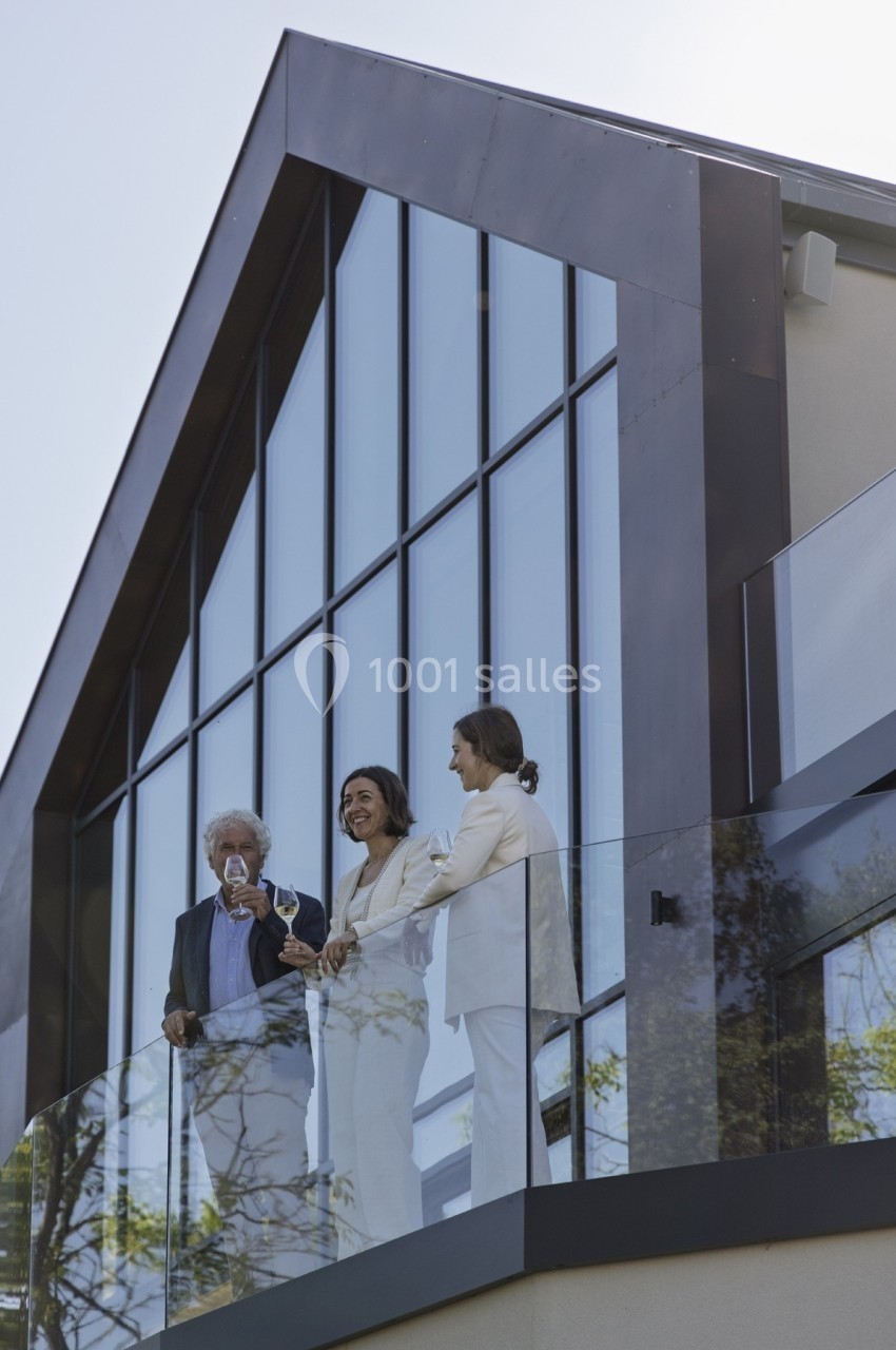 Trois personnes debout sur un balcon vitré devant une façade moderne avec de grandes fenêtres.