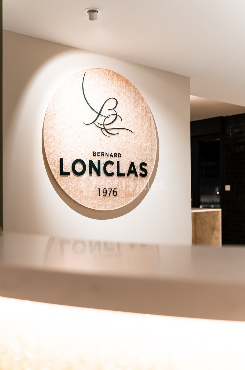 Panneau rond en bois clair avec le logo et le texte ’Bernard Lonclas 1976’ sur un mur intérieur lumineux.