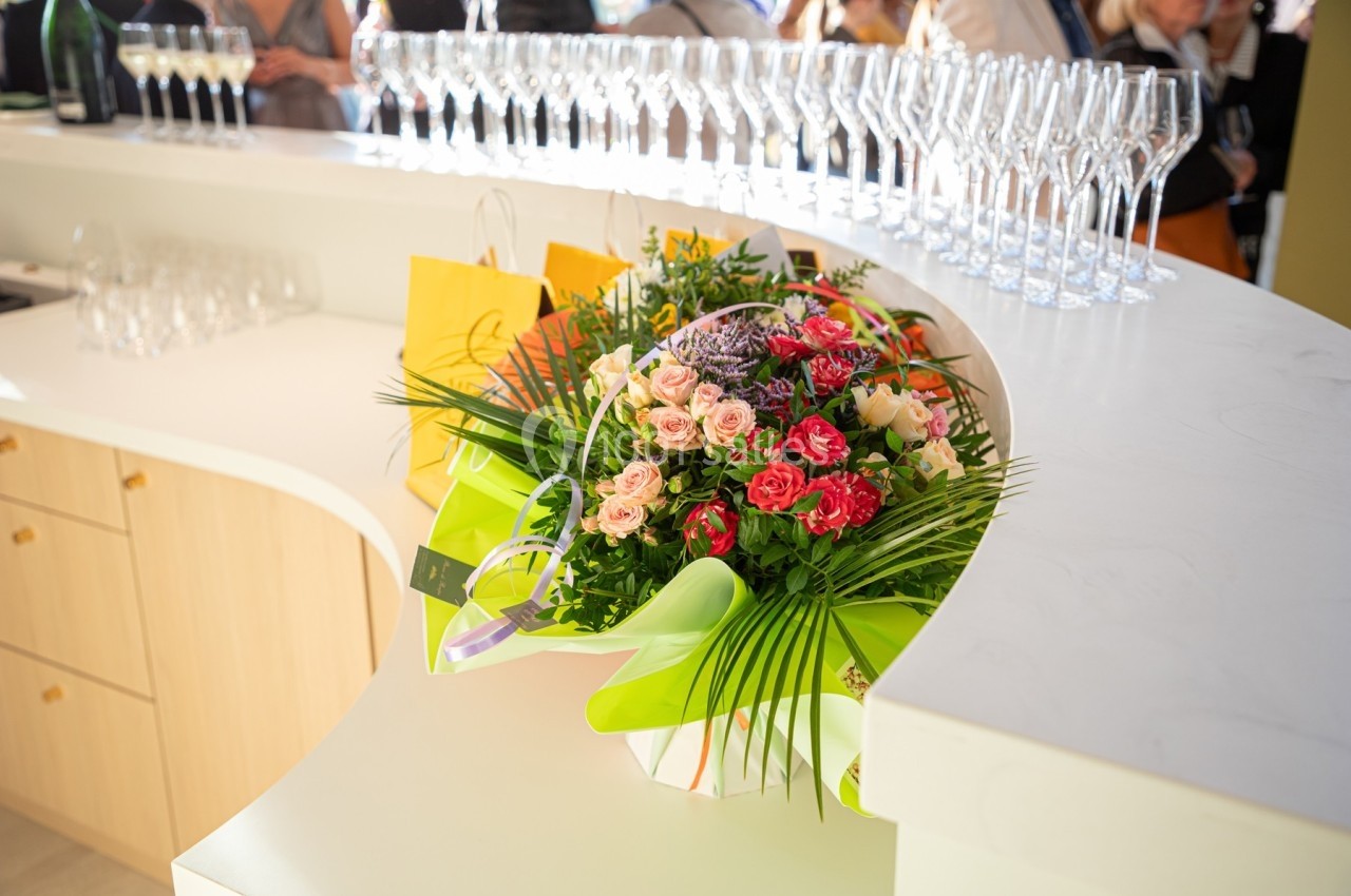 Bouquet de fleurs colorées posé sur un comptoir blanc, entouré de verres à pied et de sacs cadeaux jaunes.