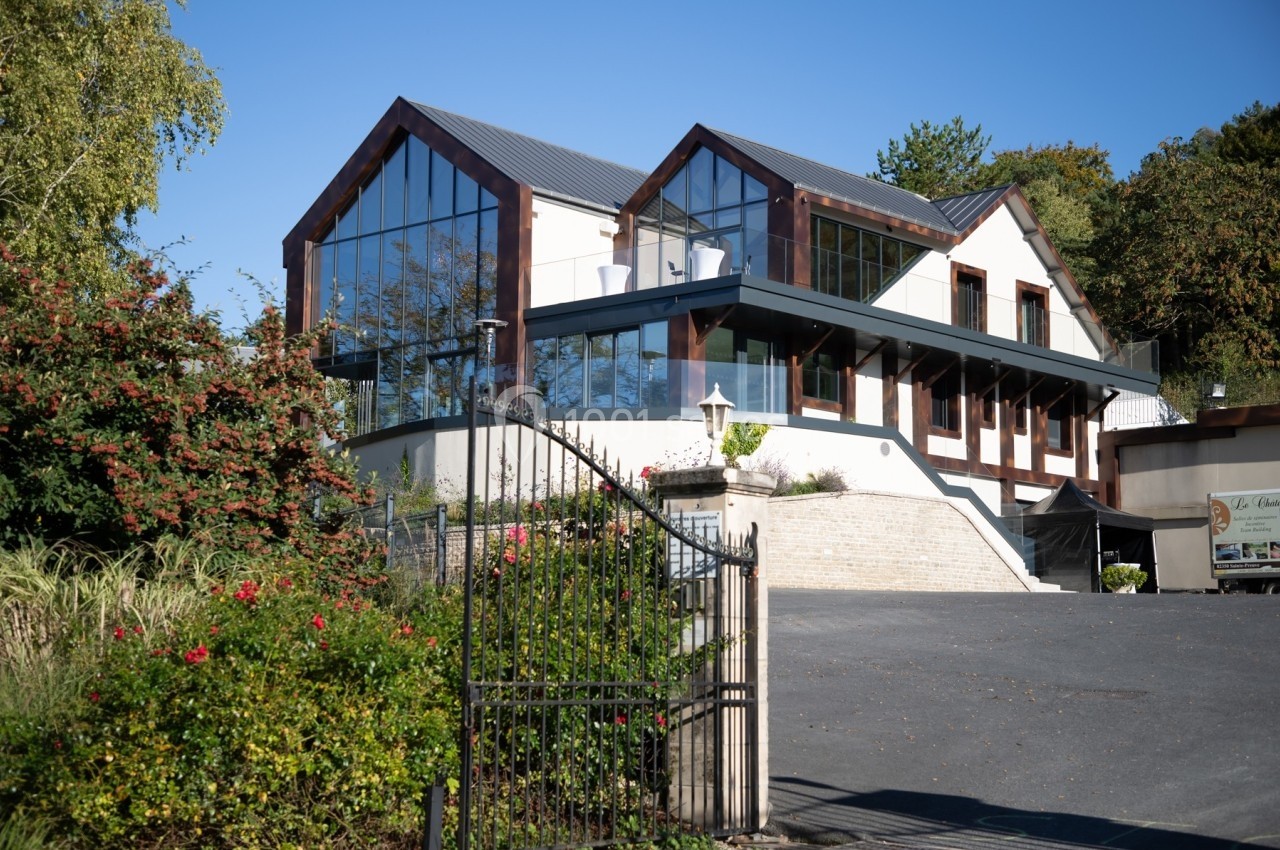 Bâtiment moderne avec façade vitrée, entouré de végétation, vu depuis une entrée avec portail en fer forgé.