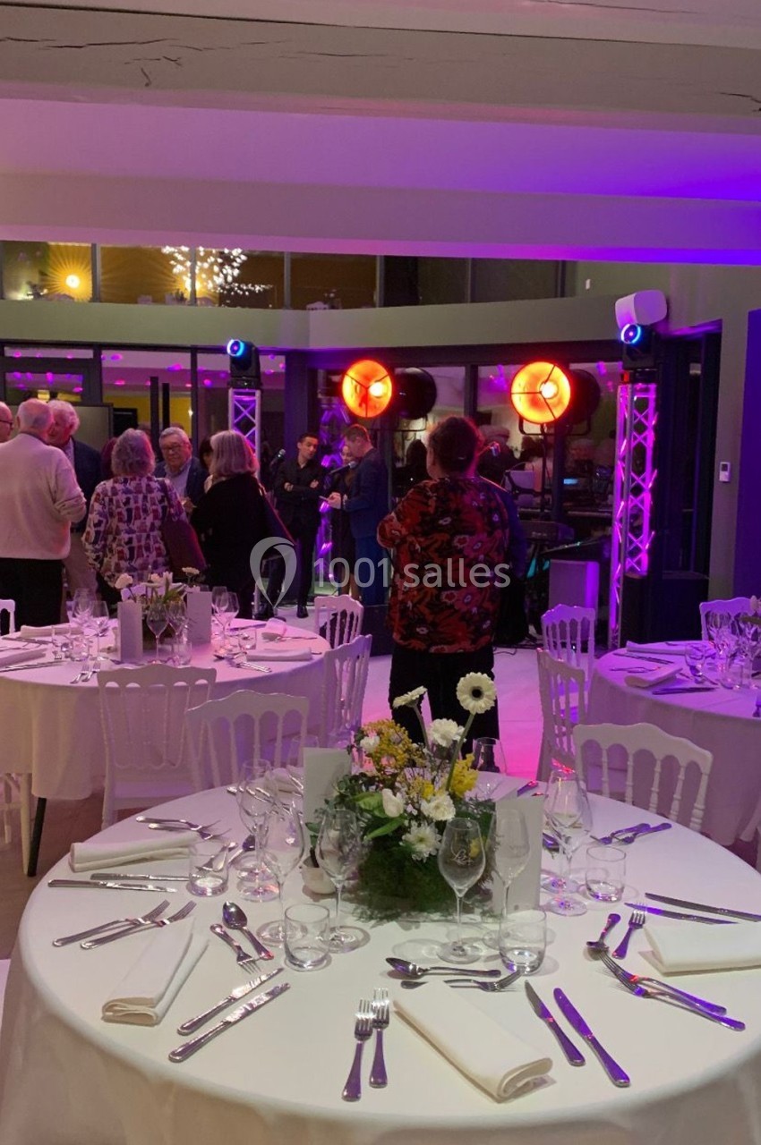 Salle de réception décorée avec des tables dressées, des invités debout discutant près d'un espace éclairé.