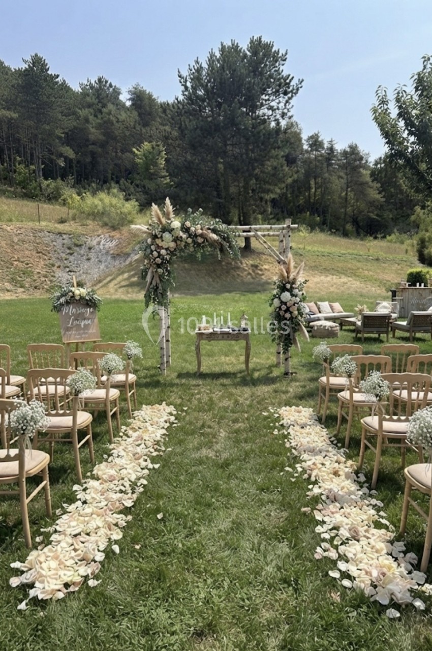 Allée décorée de pétales menant à une arche fleurie pour une cérémonie en plein air, entourée de chaises en bois.