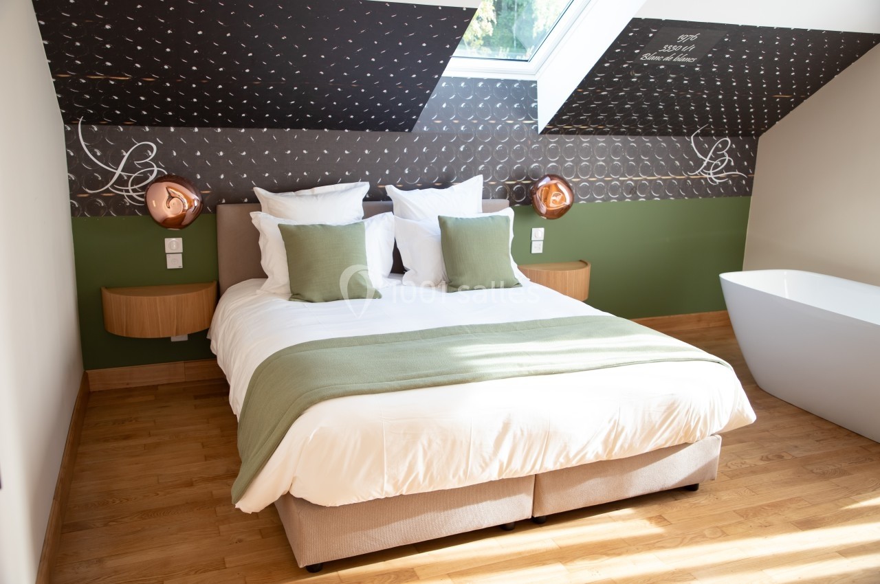 Chambre lumineuse sous combles avec lit double, couvre-lit vert, deux lampes murales et une baignoire blanche.